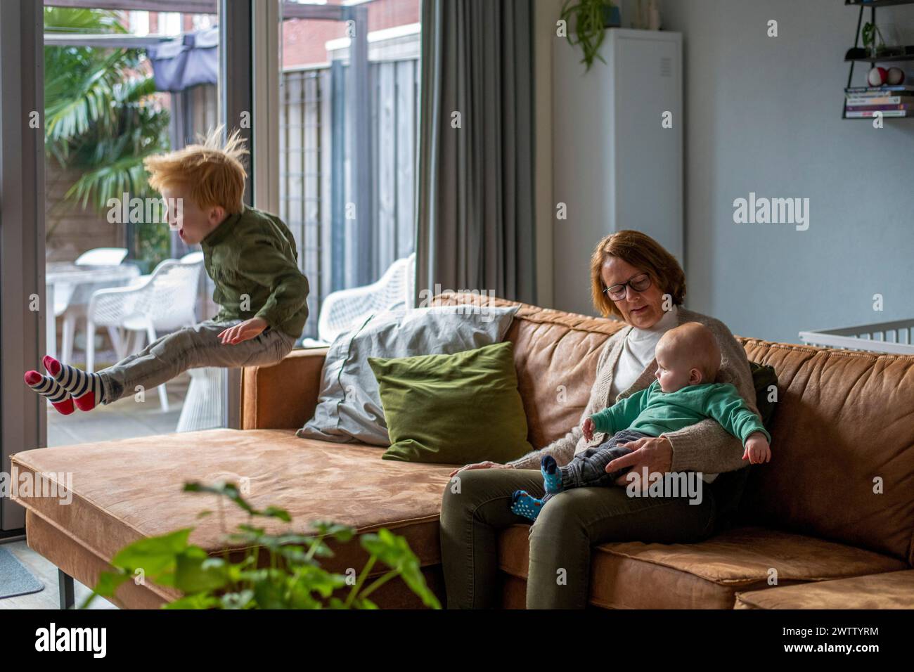 Familienmoment mit einem Kind, das auf der Couch springt, einem Kleinkind auf dem Schoß einer Frau ruht und einem gemütlichen Innenraum. Stockfoto