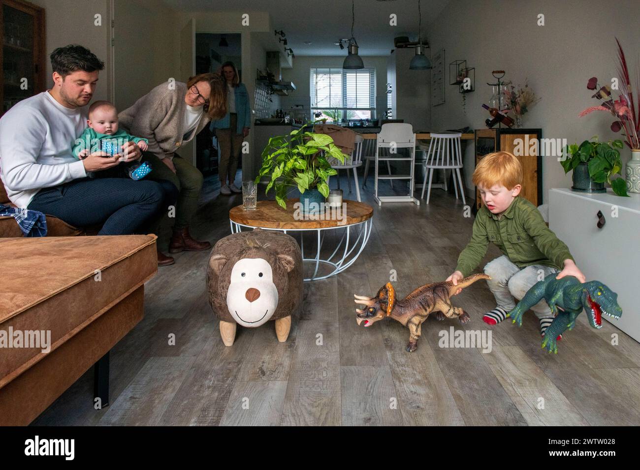 Eine Familie verbringt Zeit zusammen drinnen, mit einem Mann, der ein Baby hält, einer Frau, die das Kind ansieht, und einem kleinen Jungen, der mit Dinosaurierspielzeug auf dem Boden spielt. Stockfoto