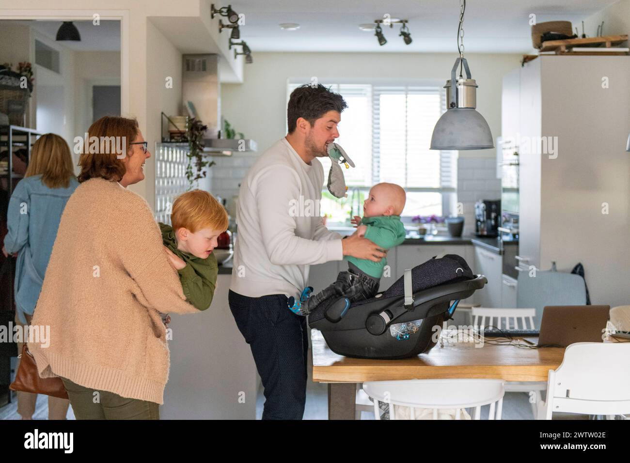 Ein Familienmoment in einer gemütlichen Küche, während ein Vater spielerisch ein Baby aus einem Autositz hebt, während ein Kind und eine Großmutter zuschauen. Stockfoto