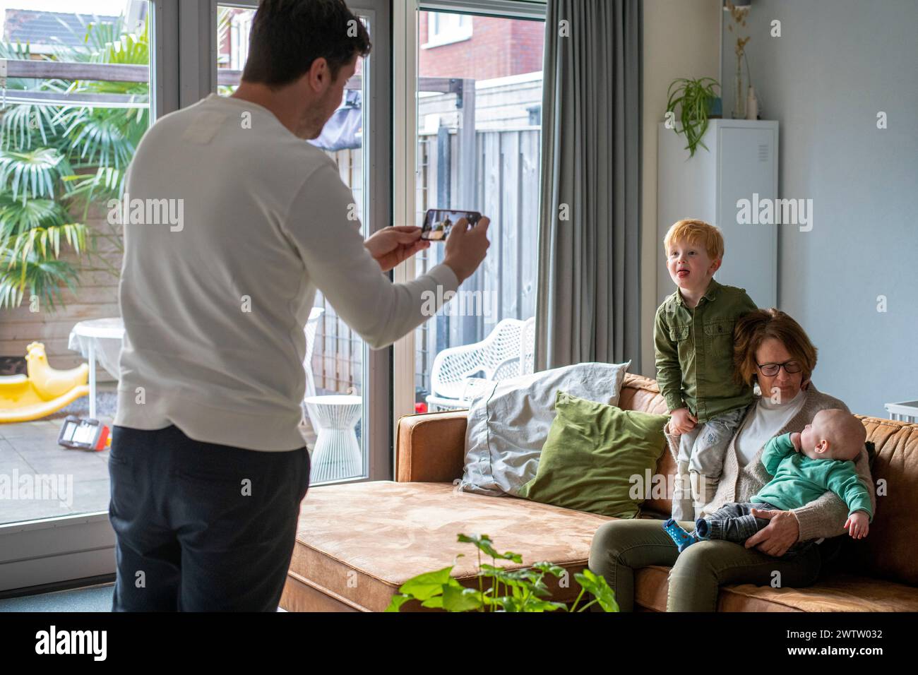 Ein Familienmoment, der drinnen festgehalten wird, während ein Mann ein Foto von einer Frau mit zwei kleinen Kindern auf einem gemütlichen Sofa macht. Stockfoto