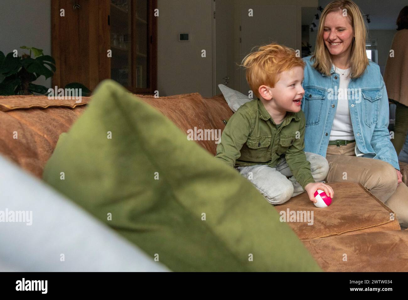 Kleines Kind lächelt vor Freude neben einer sitzenden Frau in einem gemütlichen Wohnzimmer. Stockfoto