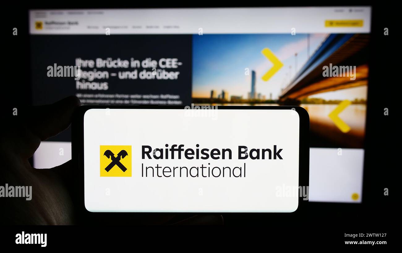 Person mit Mobiltelefon mit Logo der österreichischen Raiffeisen Bank International AG (RBI) vor der Webseite. Schwerpunkt auf dem Display des Telefons. Stockfoto