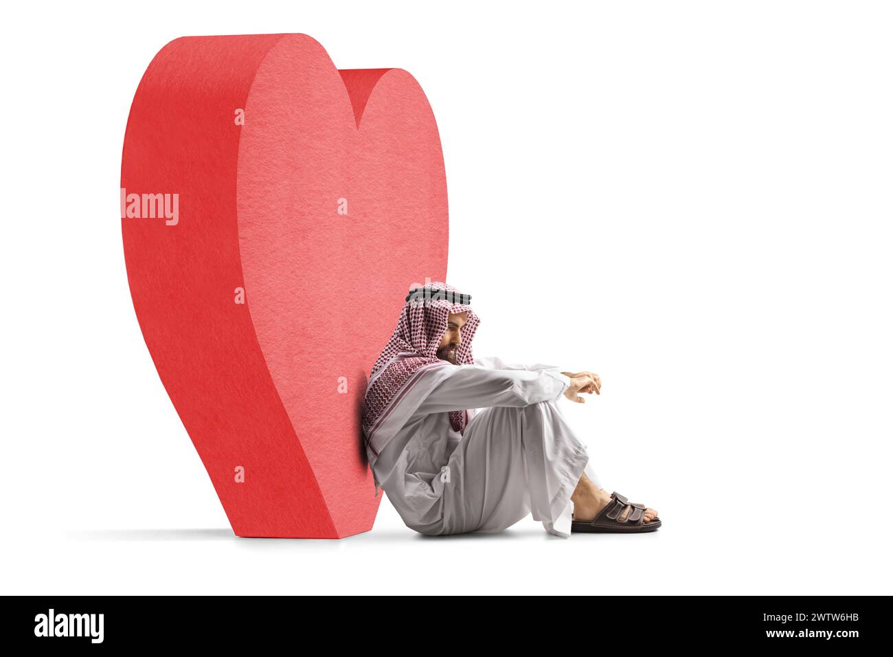 Trauriger saudi-arabischer Mann, der auf dem Boden neben einem großen roten Herzen sitzt, isoliert auf weißem Hintergrund Stockfoto