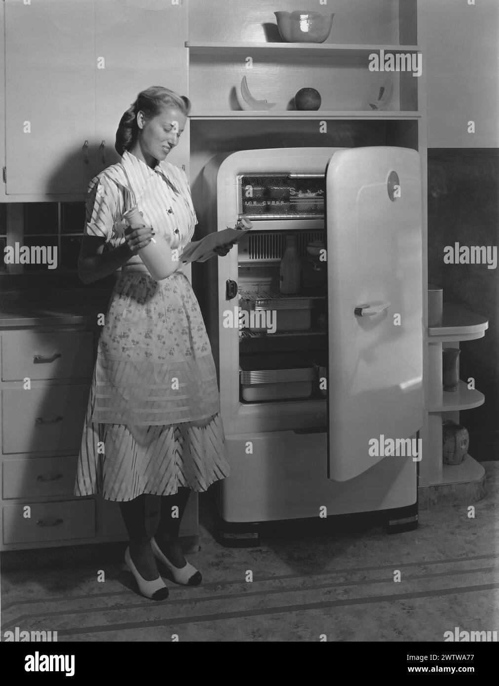 Eine junge Frau in Kleid, Schürze und Hi-Heels, die mit offener Kühlschranktür in der Küche steht, während sie ein schriftliches Rezept liest und eine Karaffe Milch hält Stockfoto