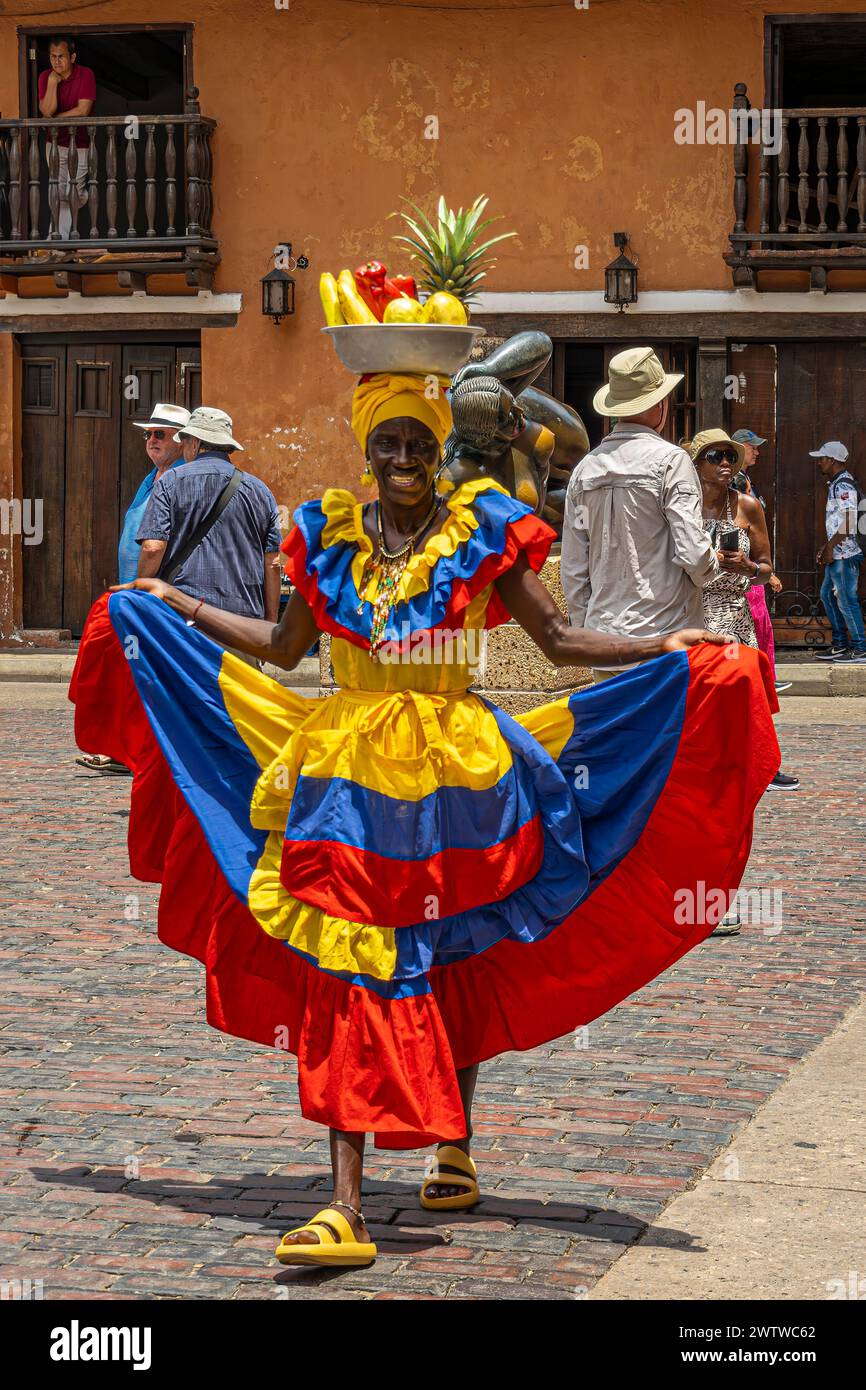 Cartagena, Kolumbien - 25. Juli 2023: Frau mit farbenfroher traditioneller Kleidung in kolumbianischen Nationalfarben tanzt auf San mit einem mit Früchten gefüllten Gericht auf dem Kopf Stockfoto