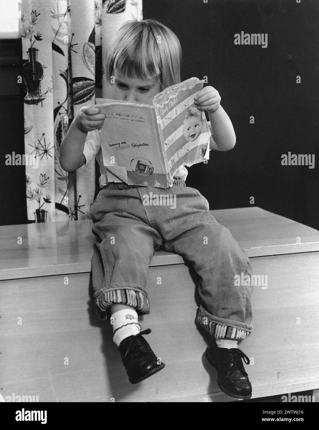 Kleiner Junge in Jeans und einer Schüssel Haarschnitt, der auf einer Holzkiste sitzt und ein Buch mit dem Titel „das erste Jahr des Lebens“ liest Stockfoto