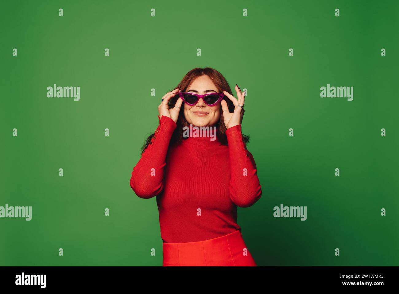Porträt einer glücklichen, stilvollen Frau, die in einem Studio mit grünem Hintergrund steht und trendige Freizeitkleidung mit modischer Sonnenbrille trägt. Ihr pulsierendes Auto Stockfoto