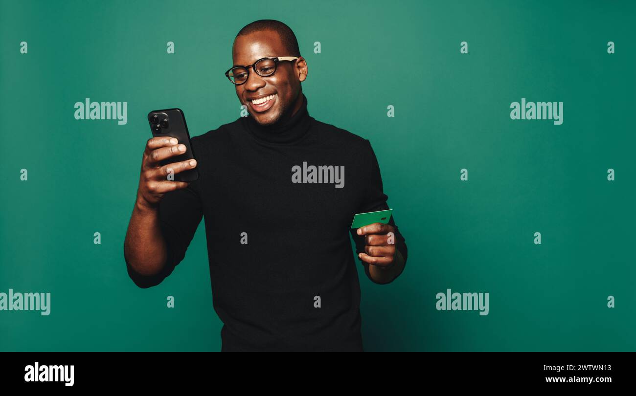Ein glücklicher junger Mann in trendiger Freizeitkleidung und Brille steht vor einem leuchtend grünen Hintergrund. Halten Sie eine elegante grüne Kreditkarte und ein Smartphone Stockfoto