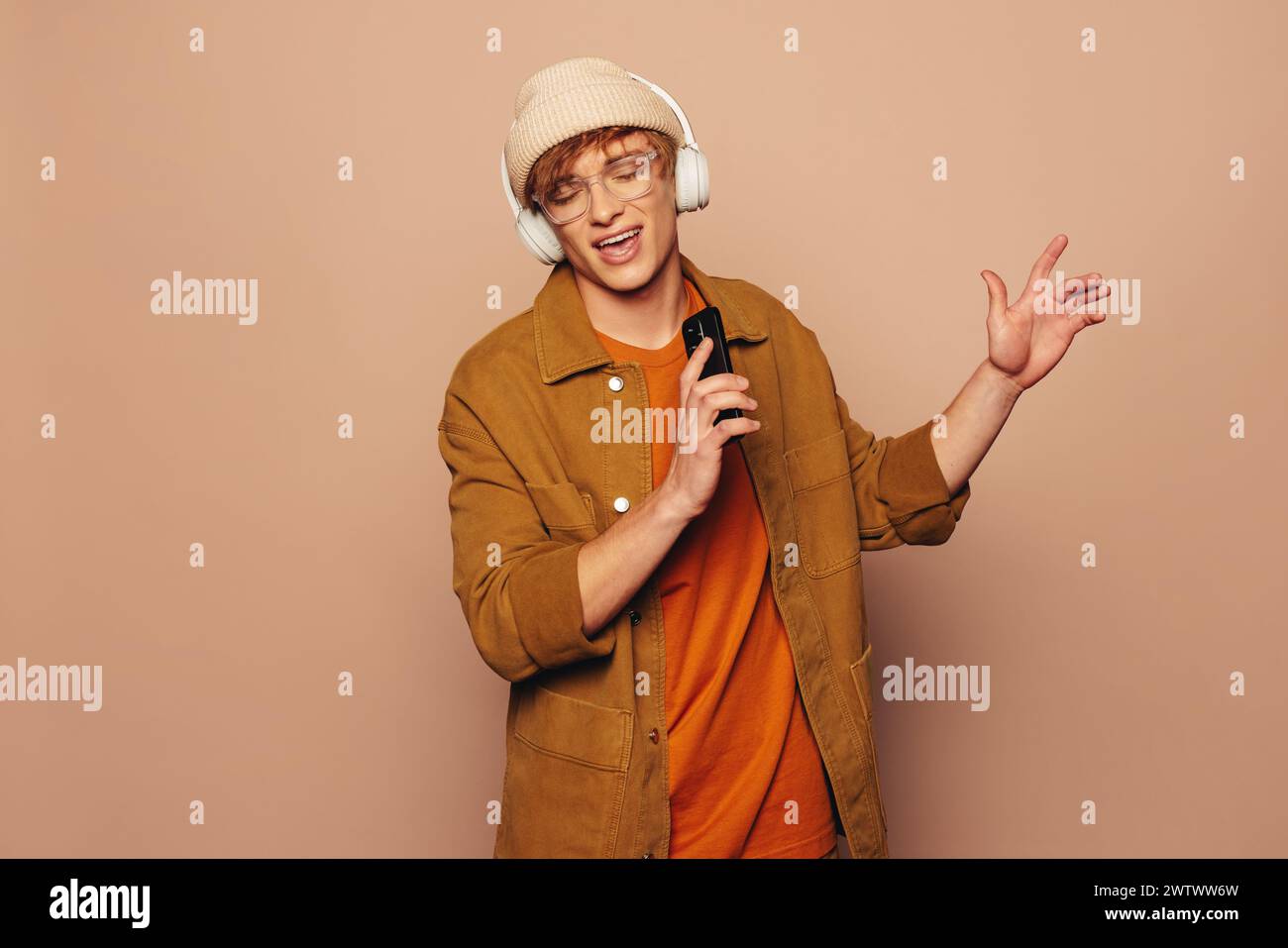 Der modische junge Mann steht vor einem lebhaften pfirsichfarbenen Hintergrund, hält ein Smartphone und trägt legere Kleidung. Mit Kopfhörern singt er mit Stockfoto