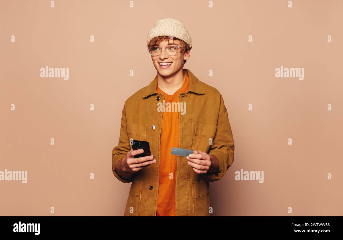 Ein lächelnder Mann in lässiger Jeansjacke nutzt sein Handy und seine Kreditkarte für Online-Banking auf einem lebhaften pfirsichfarbenen Hintergrund. Stockfoto