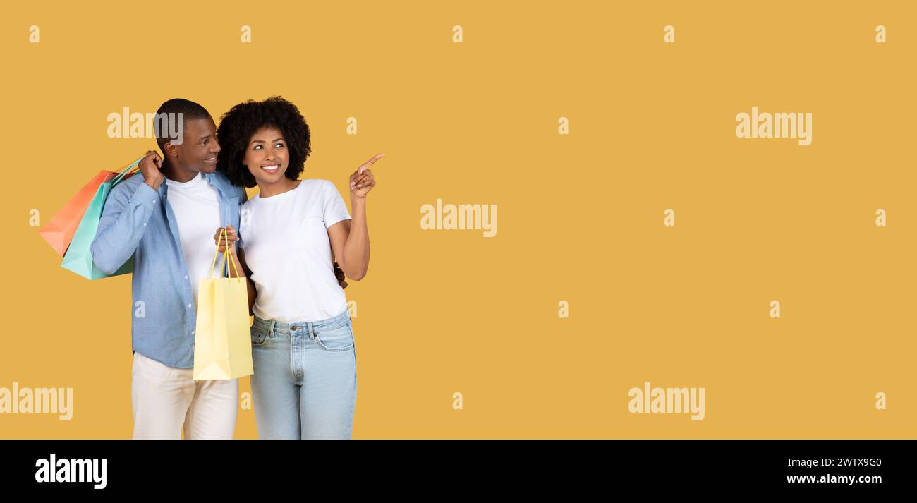 Ein entzückendes junges Paar, das bunte Einkaufstaschen hält und auf einen großen gelben Hintergrund zeigt Stockfoto