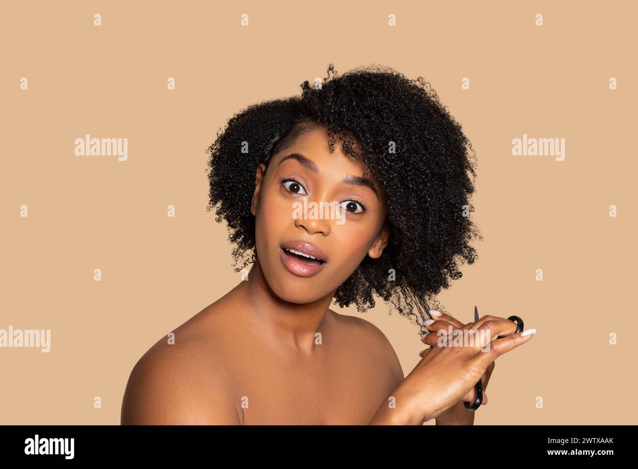 Überraschte schwarze Frau, die lockiges Haar auf beigefarbenem Hintergrund schneidet Stockfoto