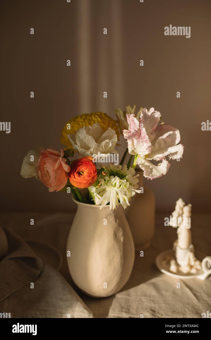 Blumenstrauß von Gerbera, Ranunkeln und Tulpenblüten in einer Vase auf einem Tisch mit einer Kerze Stockfoto