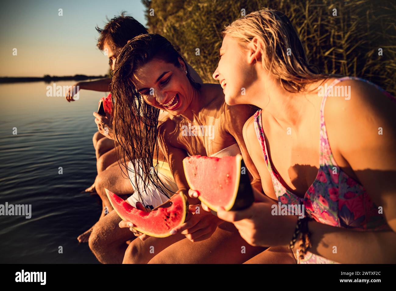 Freunde, die am Rande eines Piers sitzen und an einem Sommertag am See Wassermelonen essen. Stockfoto
