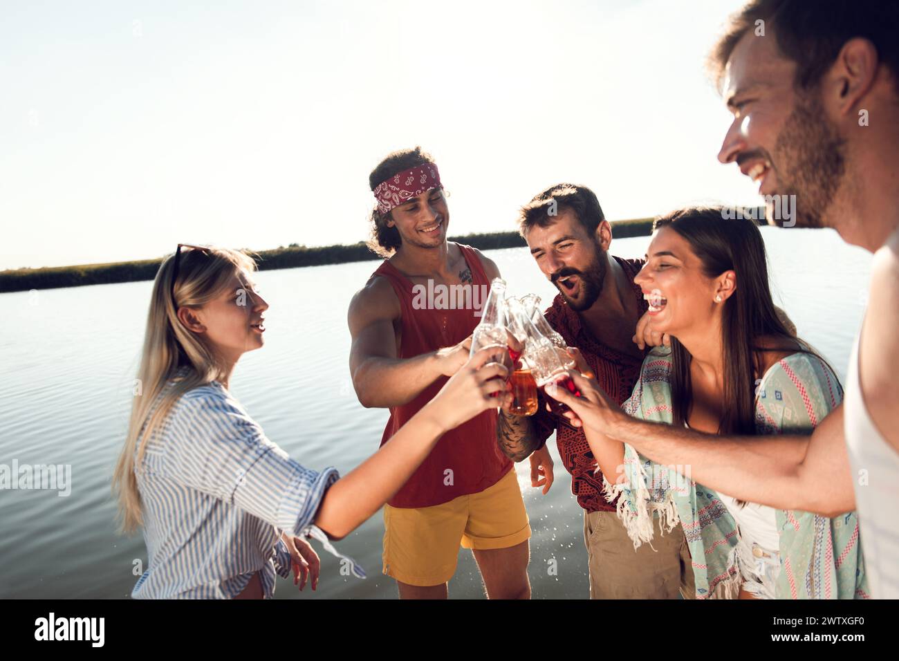 Porträt von Freunden, die auf einem Pier stehen und mit alkoholischem Getränk anstoßen und einen Sommertag am See genießen. Stockfoto