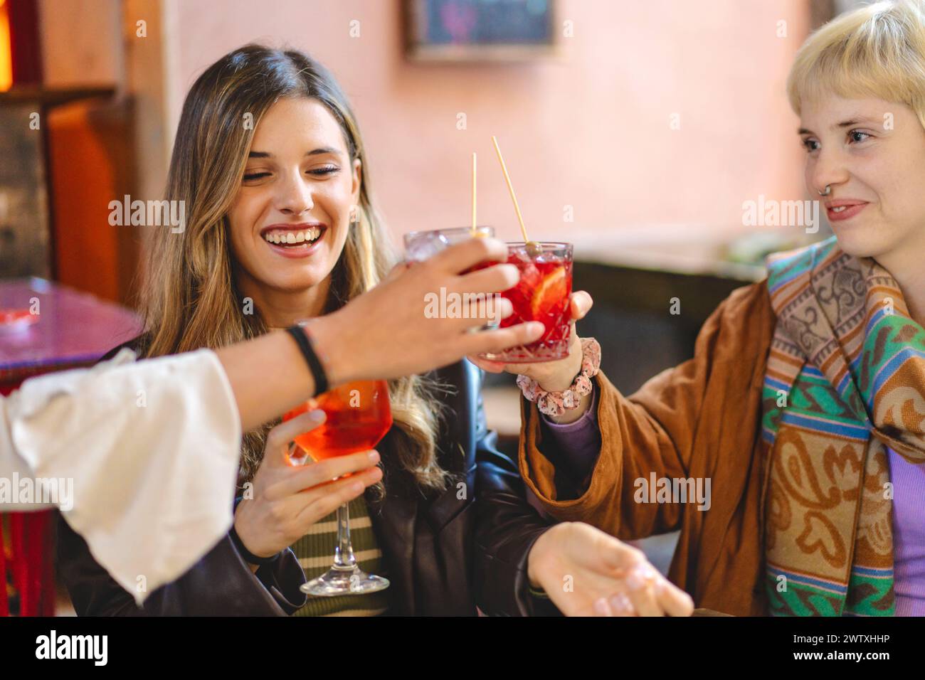 Lächelnde Freunde, die an einem gemütlichen Ort mit bunten Cocktails toben - junge Frauen feiern Freundschaft, genießen Drinks - entspannte Atmosphäre, lebhafte S Stockfoto