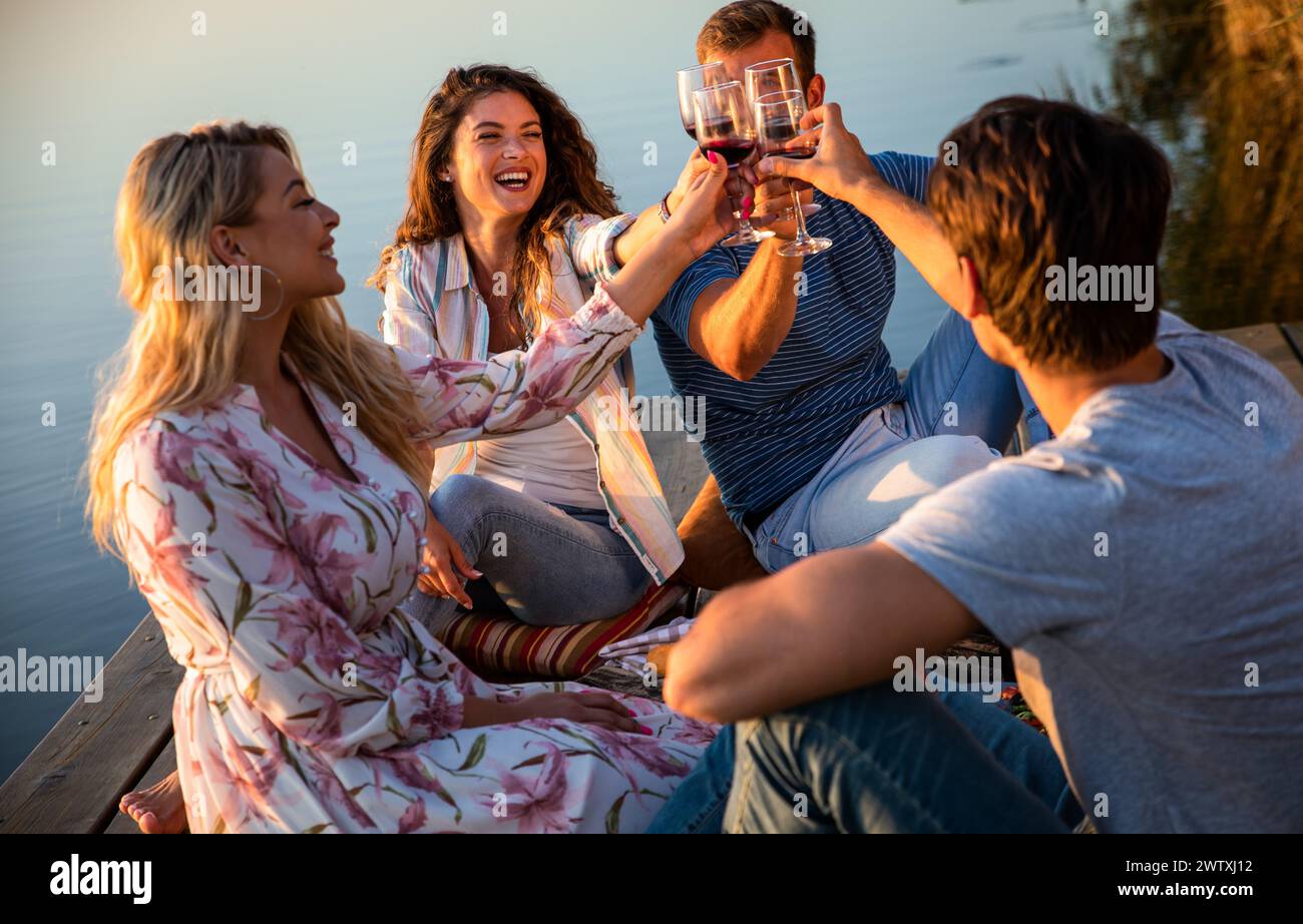 Gruppe von Freunden, die Spaß beim Picknick in der Nähe eines Sees haben, auf dem Pier sitzen und Wein trinken. Stockfoto