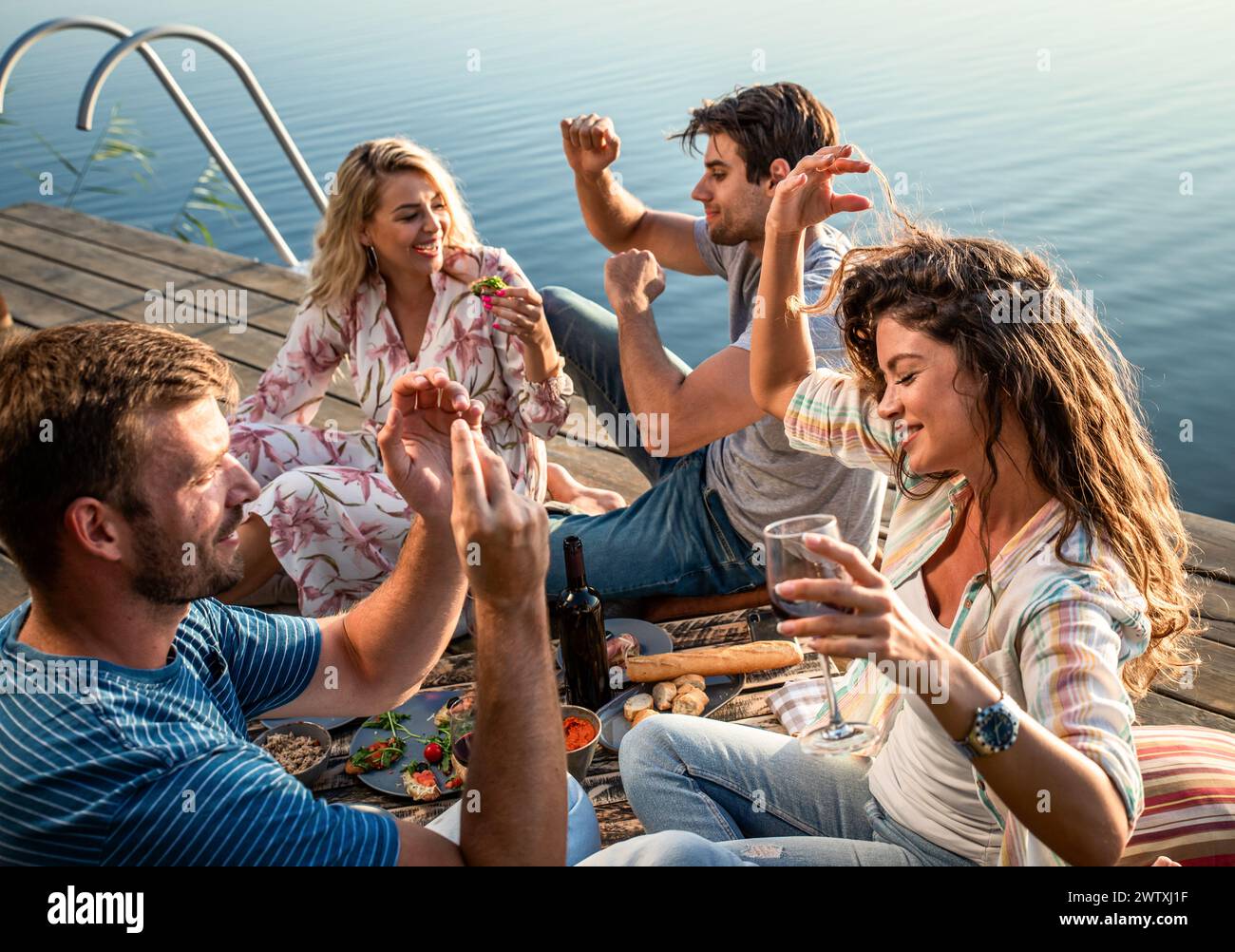 Gruppe von Freunden, die Spaß beim Picknick in der Nähe eines Sees haben, auf dem Pier sitzen und Wein trinken. Stockfoto
