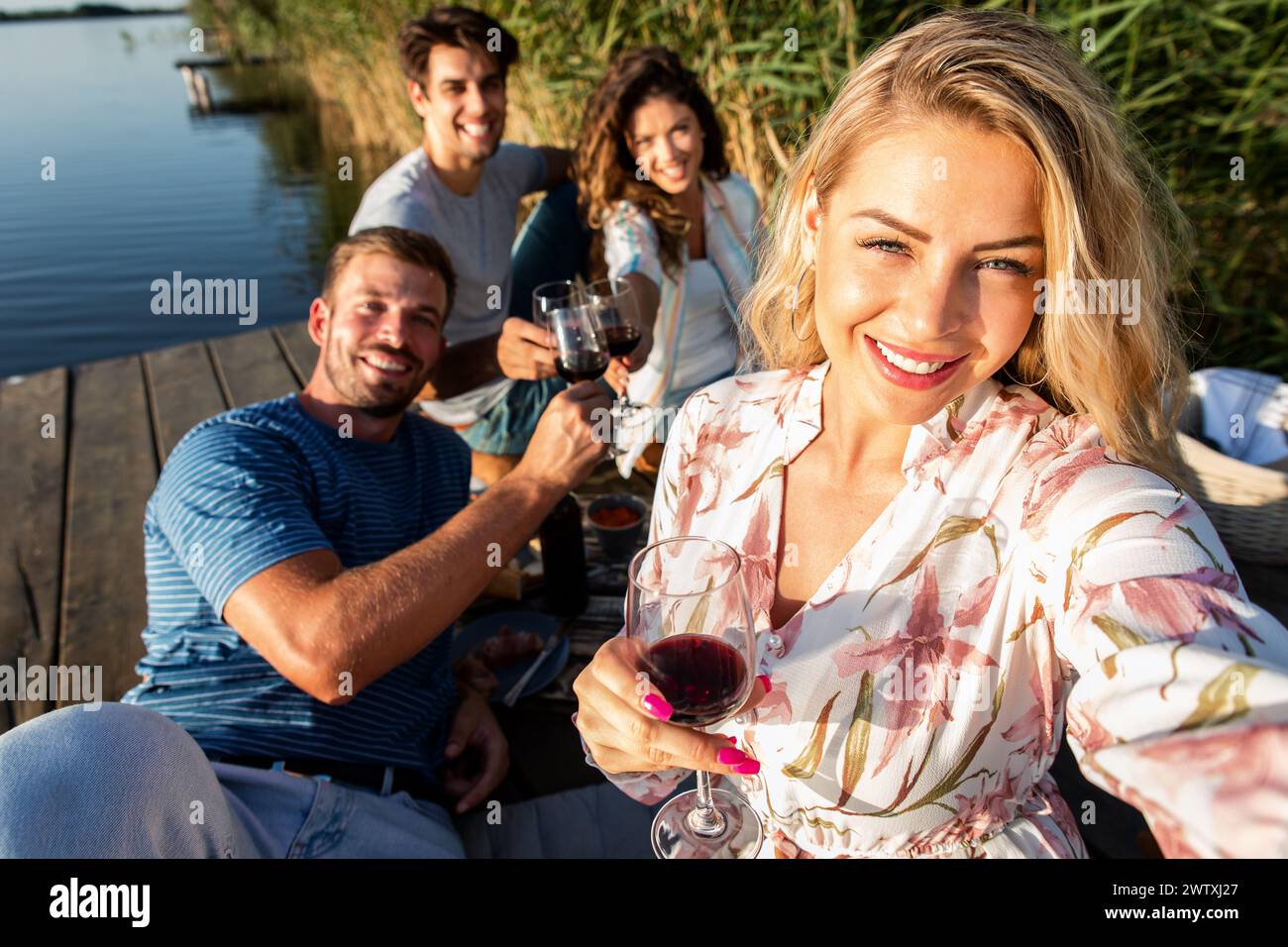 Gruppe von Freunden, die Spaß beim Picknick in der Nähe eines Sees haben, auf dem Pier sitzen und Wein trinken und Selfie machen. Stockfoto