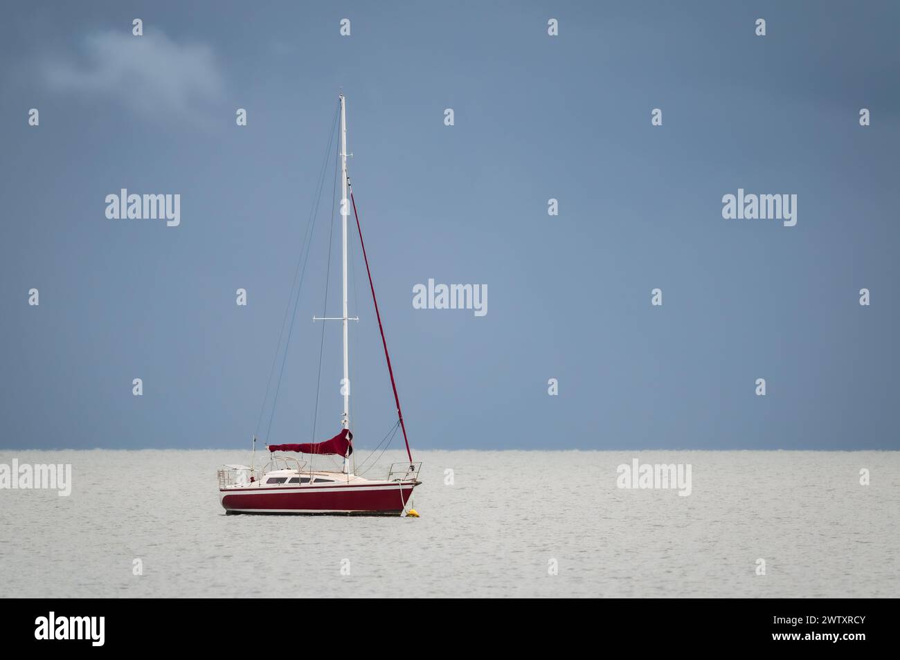 Eine einzelne, mit rotem Schalen vertäute Yacht liegt an einer ruhigen Moreton Bay mit grauem, offenem Himmel und einer einzigen weißlich-grauen Wolke. Stockfoto