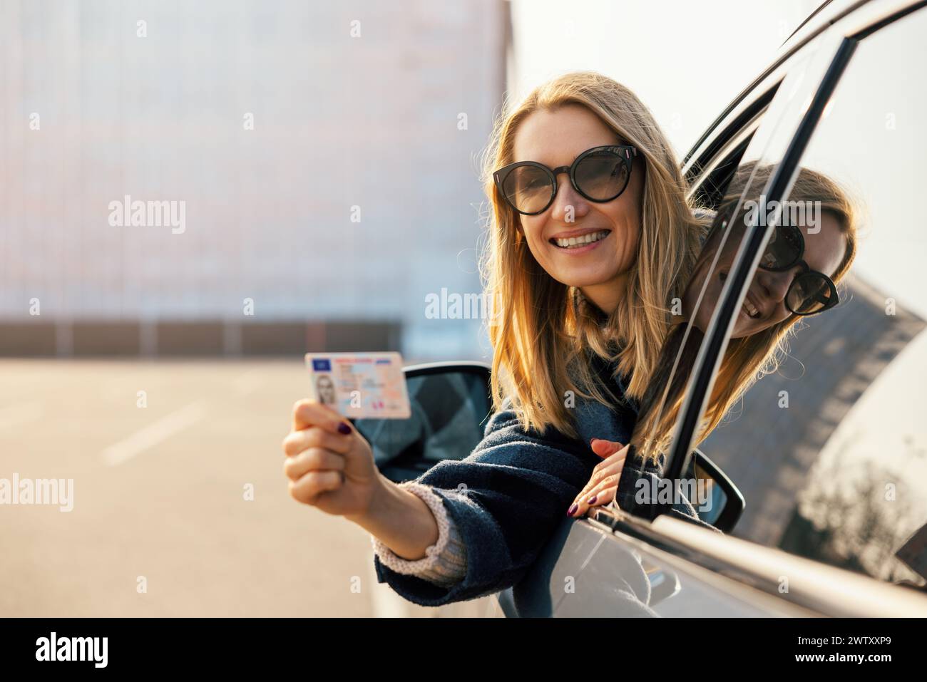 Glückliche junge Frau, die ihren neuen Führerschein nach erfolgreichem Test an der Fahrschule aus dem Autofenster zeigt. Kopierbereich Stockfoto