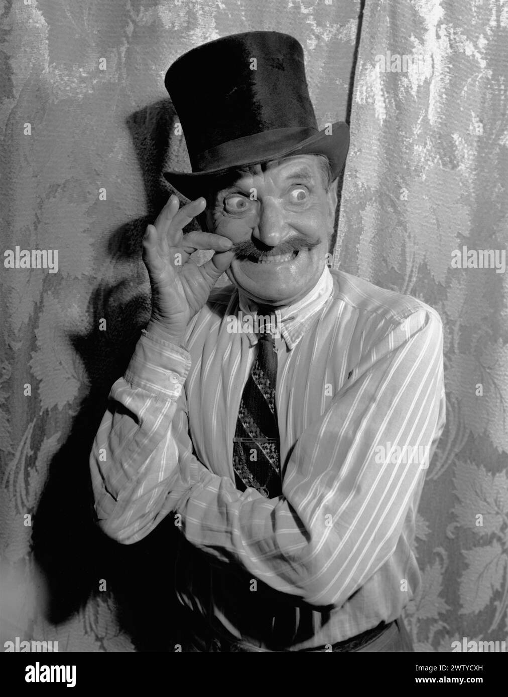 Ein älterer Mann mit Hemd und Krawatte, der einen Hut trägt, macht ein lustiges Gesicht mit prallen Augen, während er seinen Schnurrbart dreht. Um 1950 Stockfoto