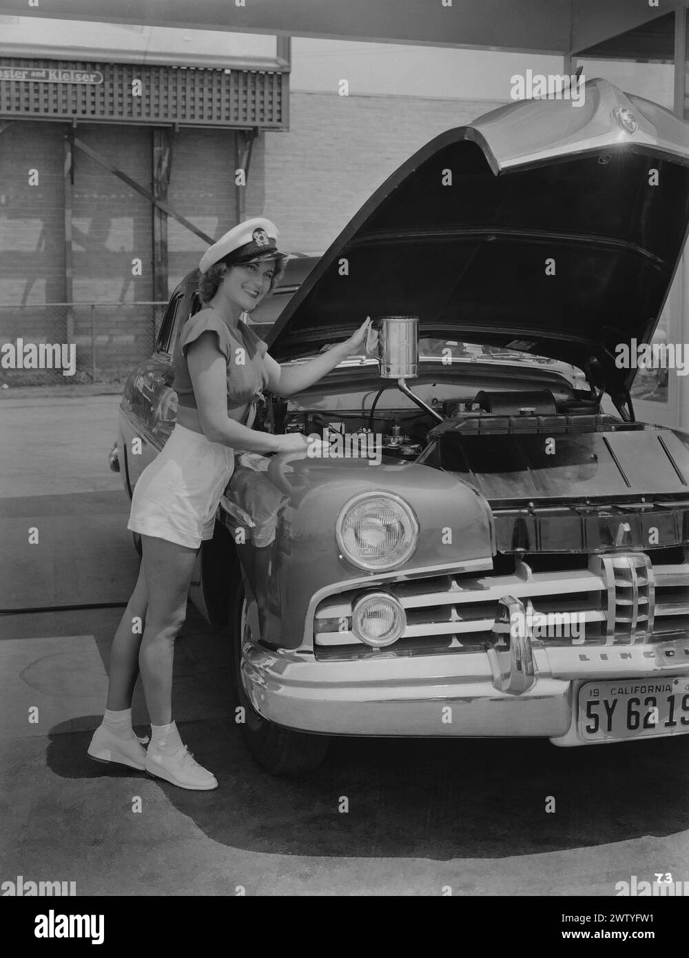 Ein Model in Shorts, Turnschuhen und einem Neckholder-Top mit Marinekappe steht neben einem Auto mit erhöhter Kapuze, die Öl auffüllt Stockfoto