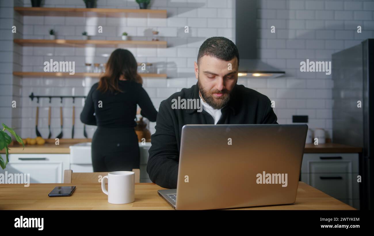 Freiberuflicher junger Mann sitzt in der Küche und arbeitet am Laptop seine Freundin kommt hinter Umarmungen herum, lächelnd, Mädchen zeigt mit dem Finger auf den Bildschirm Stockfoto