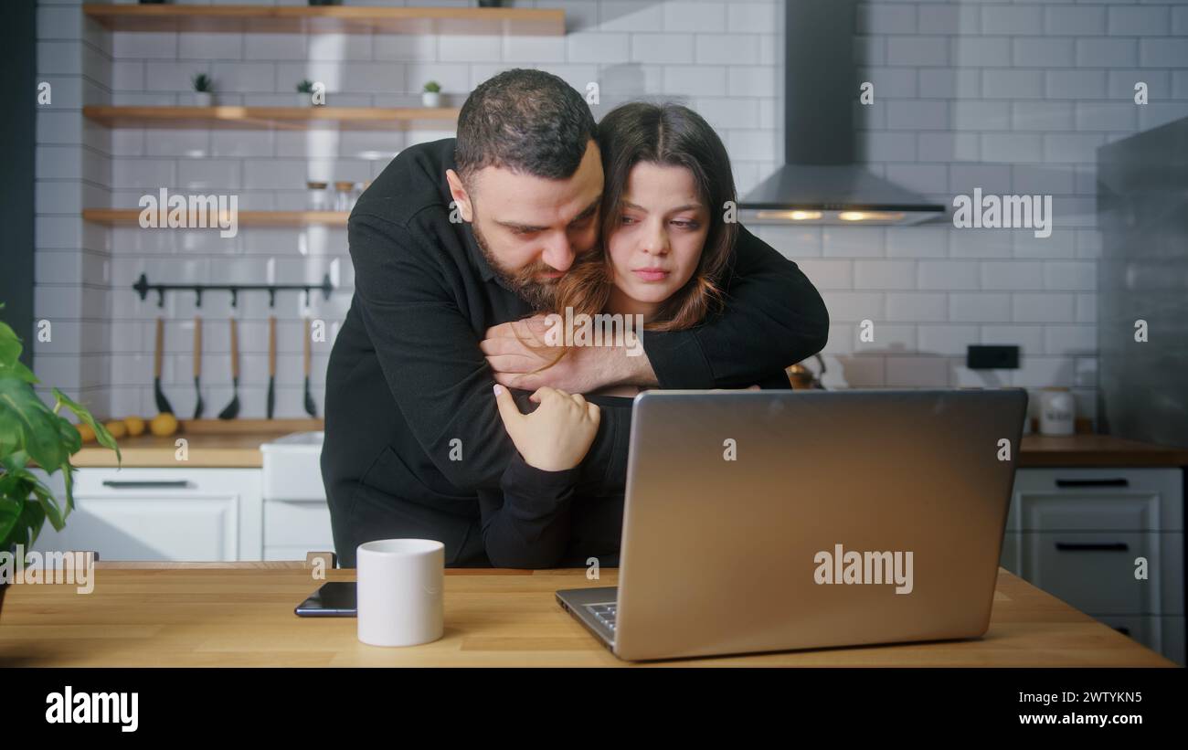 Traurige junge Frau sitzt in der Küche mit Laptop, ihr Freund umarmt sie von hinten, um sie zu trösten. Enttäuschte schlechte Nachrichten, Konzept für negatives Schadenergebnis Stockfoto