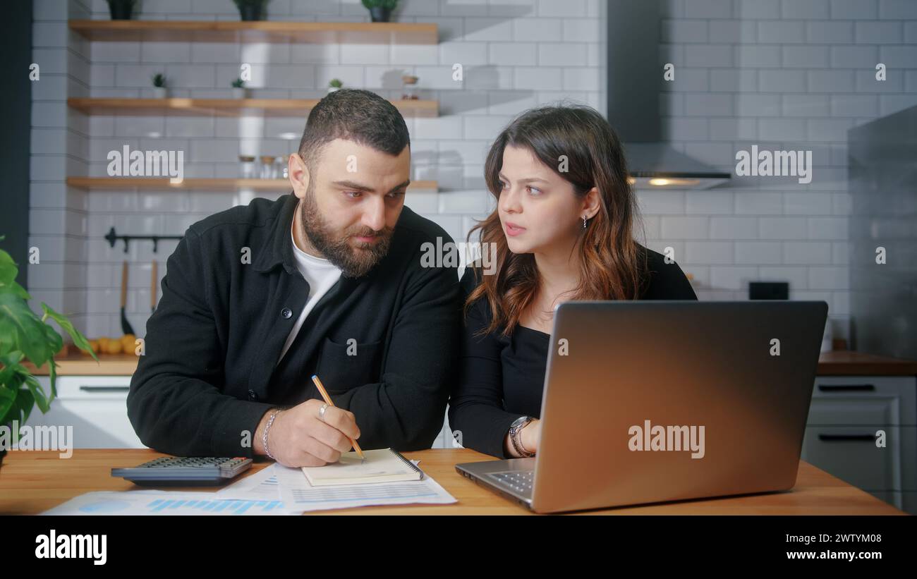 Junge Paare sitzen in der Küche und bezahlen Rechnungen mit dem Laptop, verwenden Taschenrechner Zählkosten, führen Budgetkontrolle durch, verwalten Finanzen, überprüfen überfällige Kreditzahlungen Stockfoto