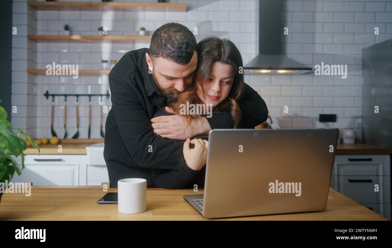 Traurige junge Frau sitzt in der Küche mit Laptop, ihr Freund umarmt sie von hinten und tröstet sie. Enttäuschte schlechte Nachrichten, Verlust negatives Ergebnis Stockfoto