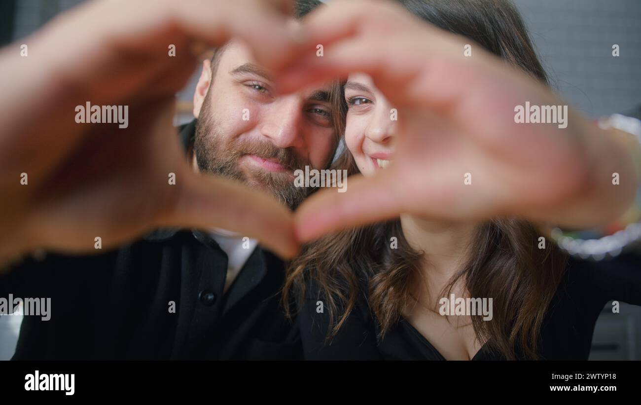 Nahaufnahme eines jungen glücklichen Paares, das mit verbundenen Fingern Herzform macht und dazwischen mit einem Lächeln in die Kamera blickt. Stockfoto