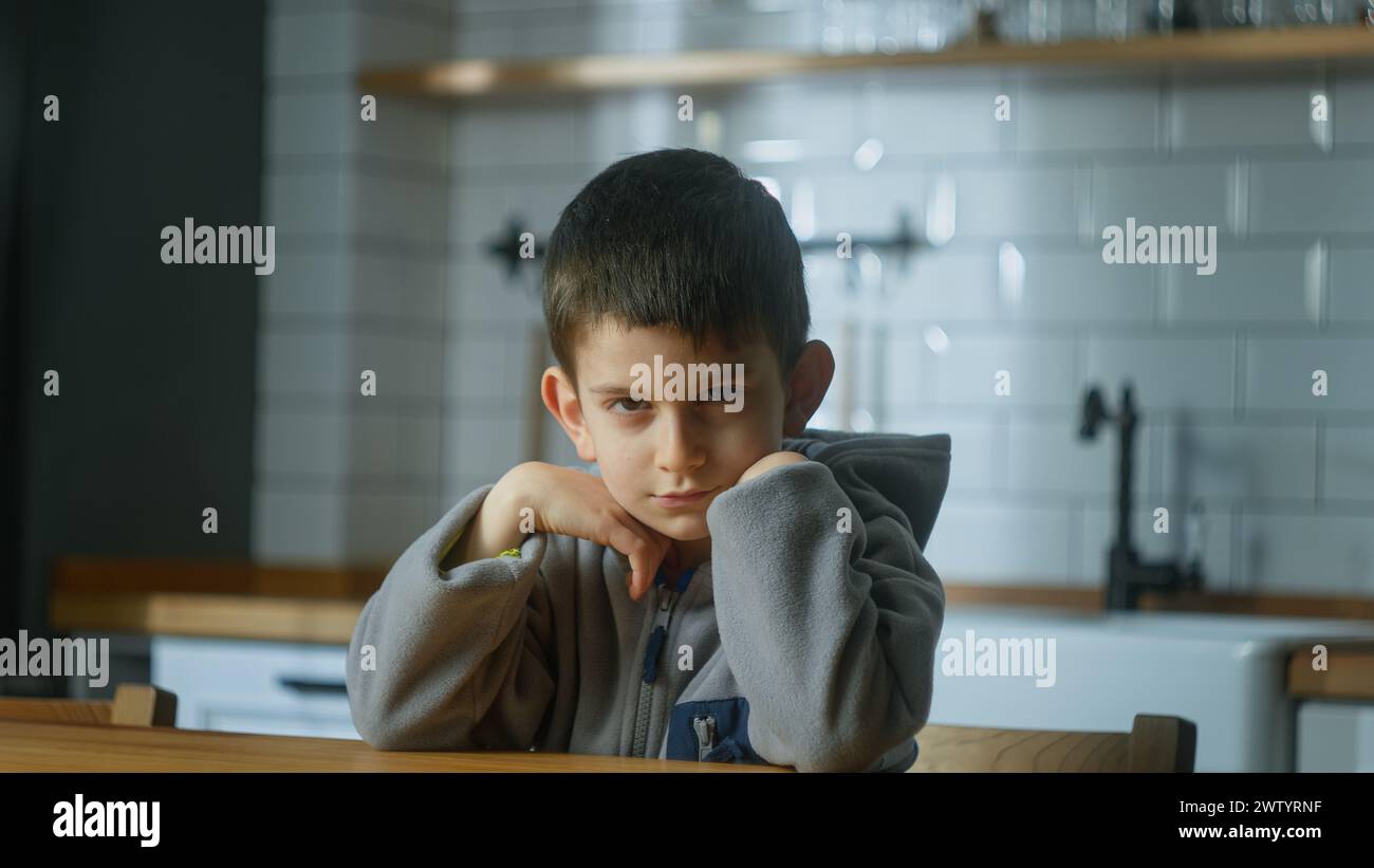 Porträt eines traurigen kleinen gekränkten Jungen, der in der Küche sitzt und in die Kamera schaut. Missverständnisse, verärgert Kind, unglücklich, unzufrieden Stockfoto