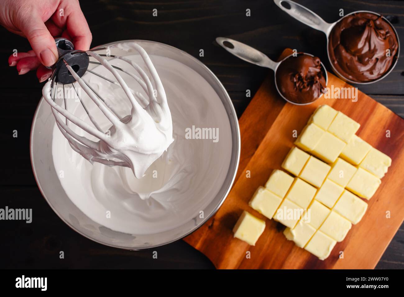 Hand hält einen Ständer Mixer Schneebesen-Aufsatz bedeckt mit Meringue: Frauenhand hält eine mit Schweizer Meringue bedeckte Drahtpeitsche über einer Metallrührschüssel Stockfoto