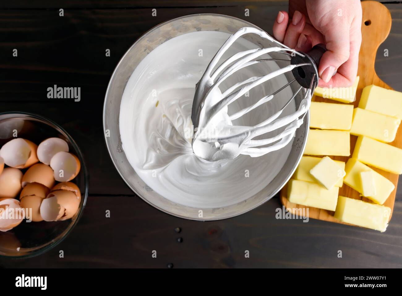 Hand hält einen Ständer Mixer Schneebesen-Aufsatz bedeckt mit Meringue: Frauenhand hält eine mit Schweizer Meringue bedeckte Drahtpeitsche über einer Metallrührschüssel Stockfoto
