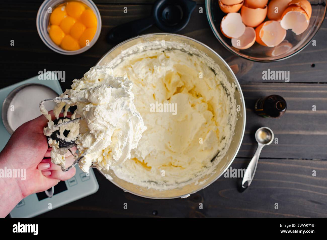 Hand hält einen Ständer Mixer Schneebesen-Aufsatz bedeckt mit Buttercreme: Frauenhand hält eine Drahtpeitsche, die mit Schweizer Meringue-Buttercreme-Frosting bedeckt ist Stockfoto