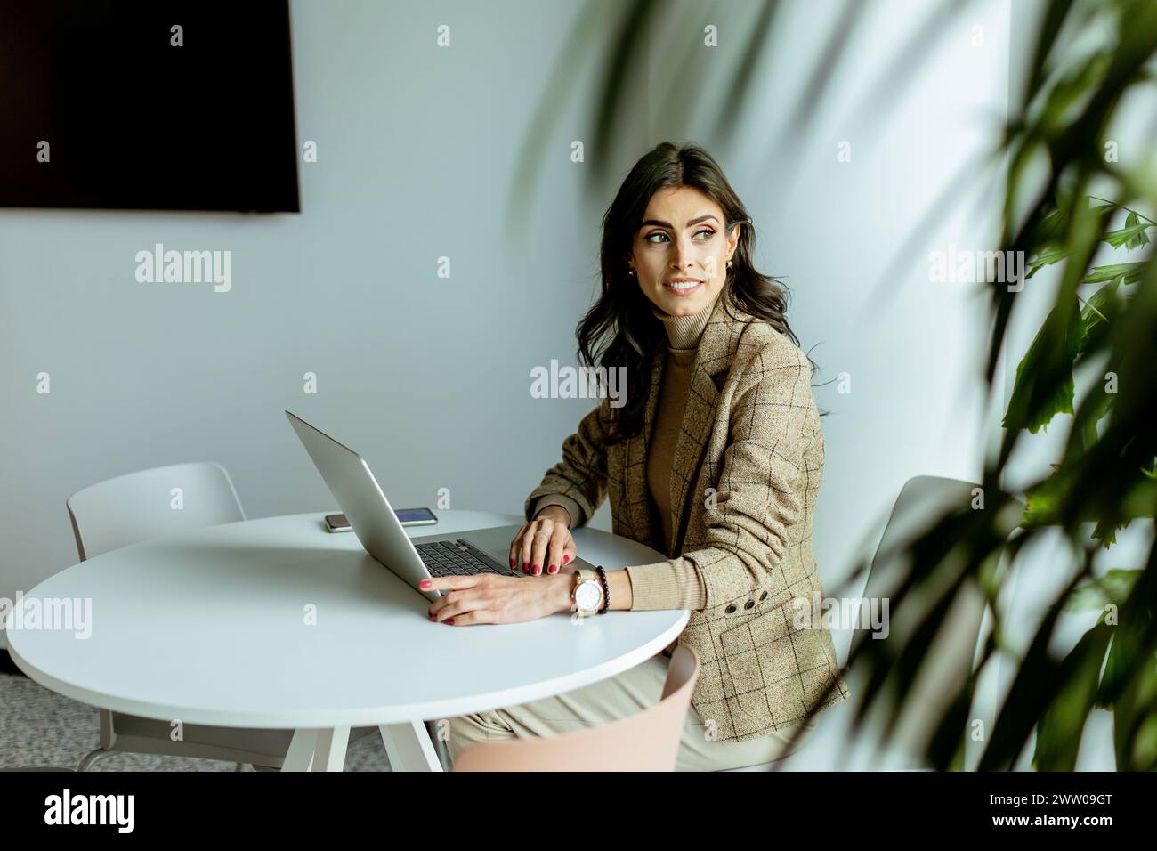 Eine konzentrierte Geschäftsfrau in einem stilvollen Blazer lässt sich auf ihrem Laptop an einem eleganten, weißen Tisch, umgeben von minimalistischem Dekor, verstecken. Stockfoto