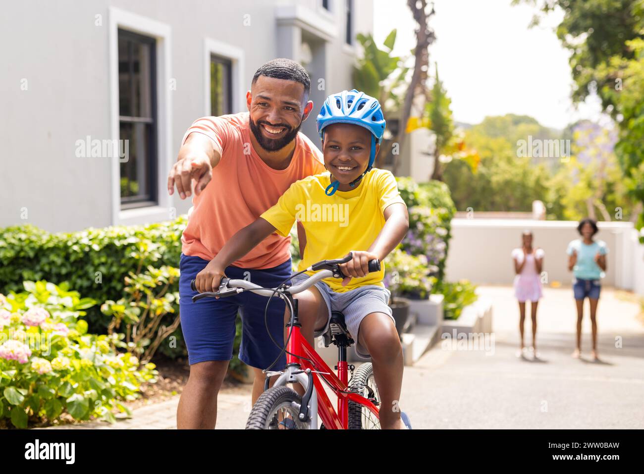 Ein afroamerikanischer Vater bringt dem Sohn bei, draußen zu Hause Fahrrad zu fahren, seine Familie beobachtet Stockfoto