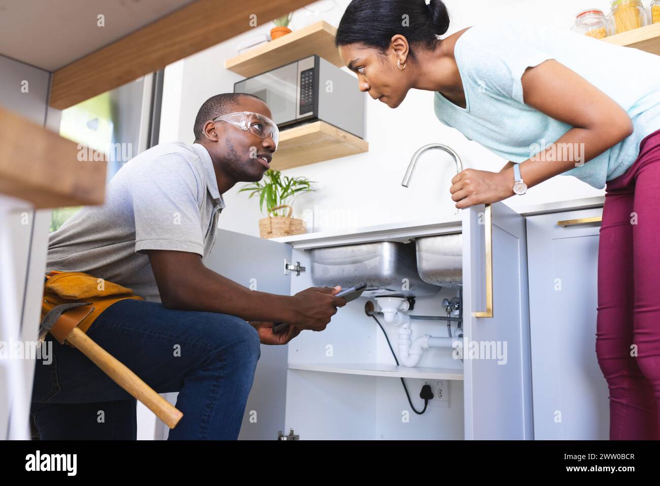 Afroamerikanischer Klempner repariert das Waschbecken, während der Hausbesitzer das Tablet benutzt Stockfoto
