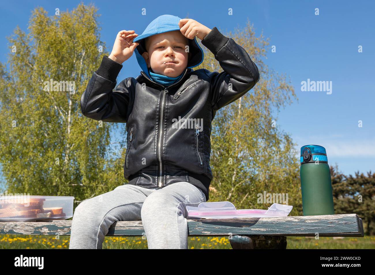 Der Junge isst in der Natur auf einer Bank zu Mittag. Guter Appetit an der frischen Luft, er steckt seinen Mund voll und sitzt mit geschwollenen Wangen. Stockfoto