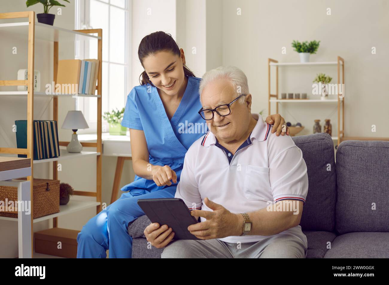 Junge hübsche brünette Krankenschwester zu Hause kommuniziert mit einem älteren Mann mit Brille. Stockfoto