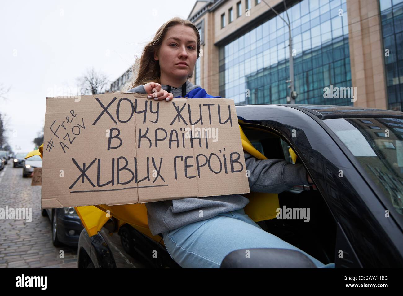 Die junge Frau zeigt ein Banner: "Ich will in Einem Land lebendiger Helden leben. Asov frei aus dem Fenster des fahrenden Autos in Kiew - 17. März 2024 Stockfoto