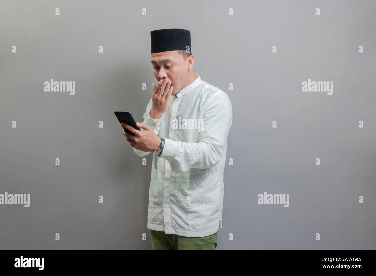 Überraschter asiatischer muslimischer Mann, der Smartphone-Handy für Anrufe, soziale Medien, mobile Anwendung online benutzte, ein koko-Shirt und Peki mit Farben trug Stockfoto