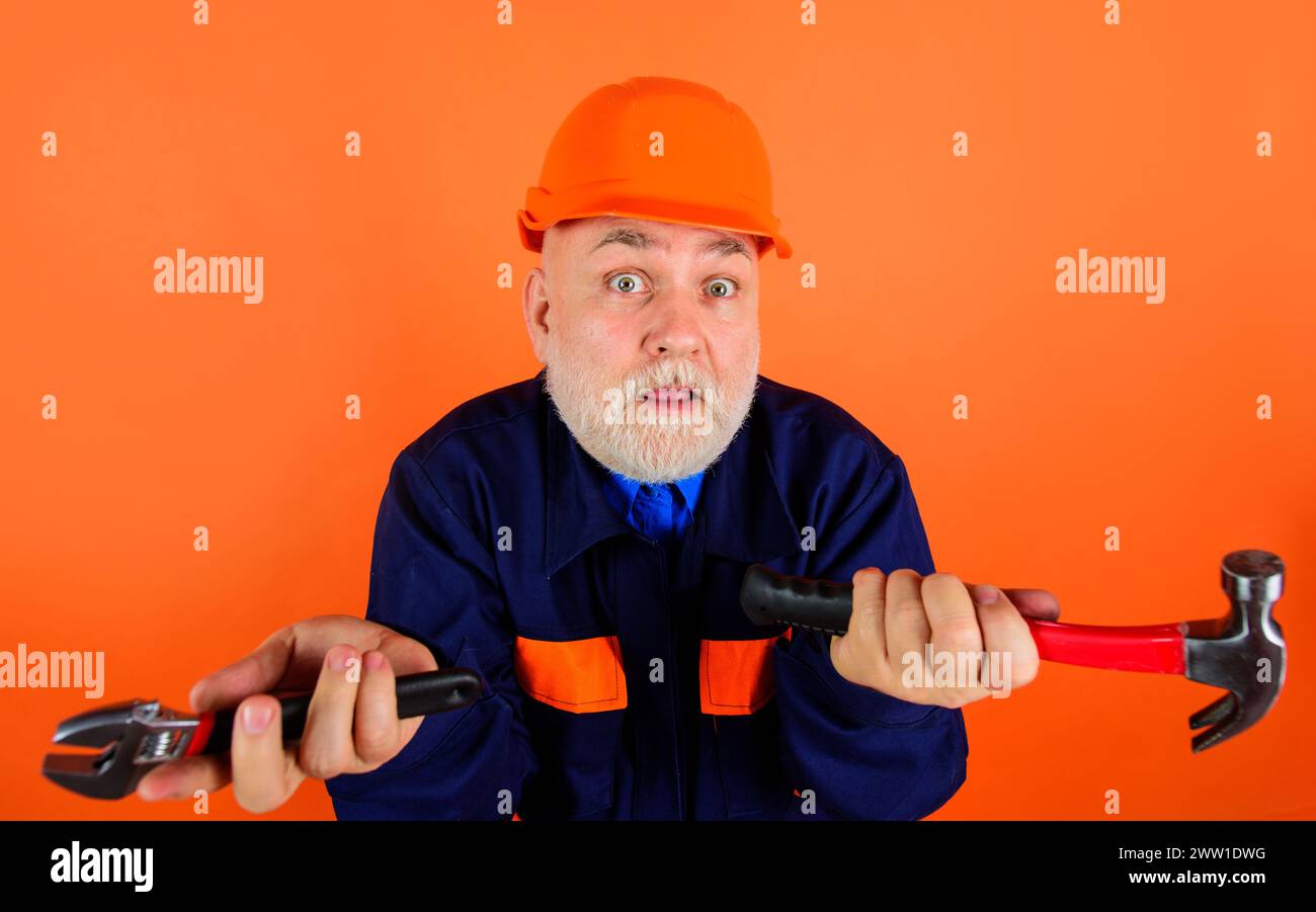 Verwirrter Erbauer im Schutzhelm mit verstellbarem Schraubenschlüssel und Hammer. Bärtiger Mechaniker oder Reparaturarbeiter in Uniform und Bauhelm mit Stockfoto