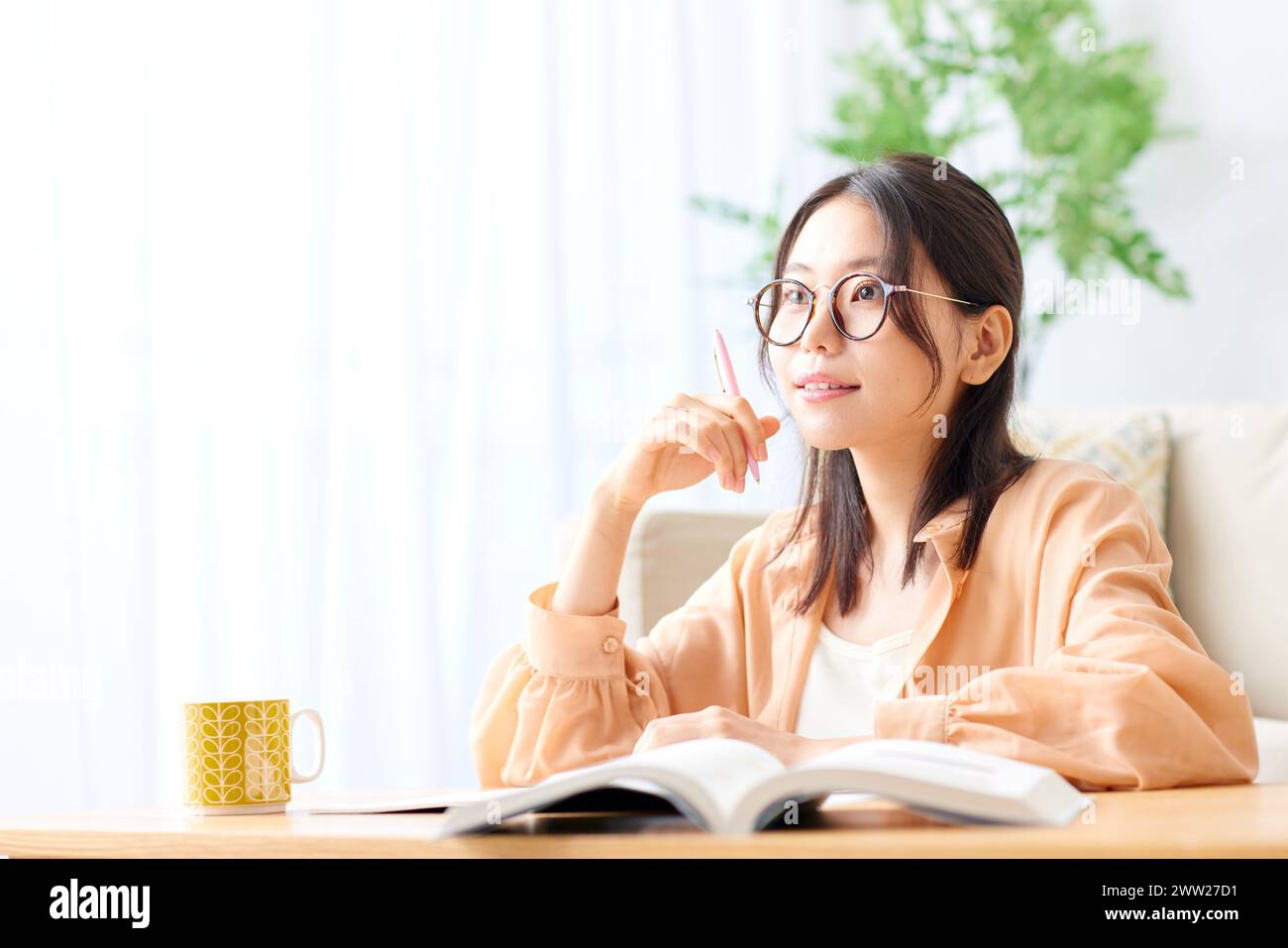 Asiatische Frau mit Brille, die mit Buch und Kaffee am Tisch sitzt Stockfoto