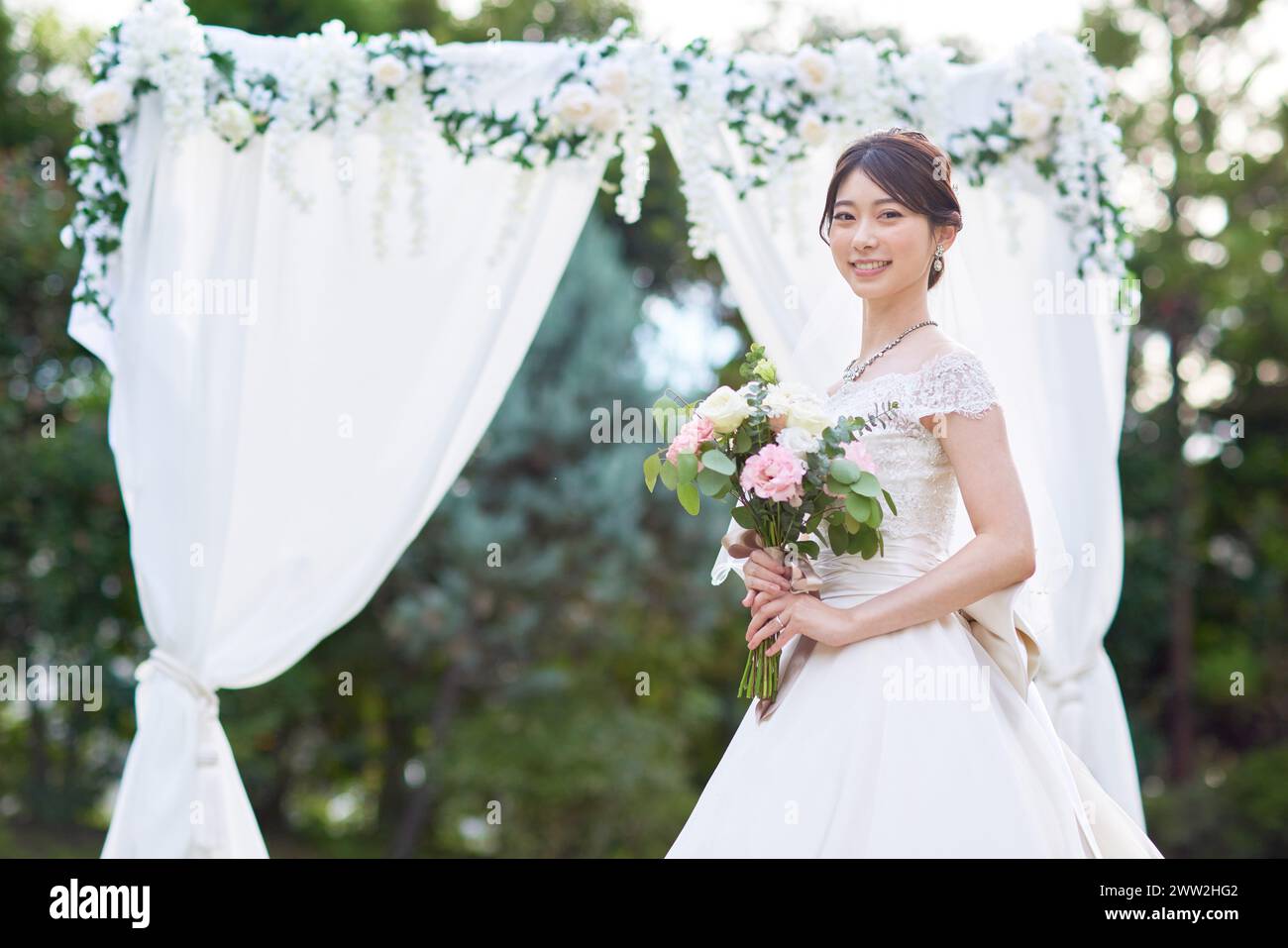 Asiatische Braut im Hochzeitskleid, die vor einem weißen Bogen steht Stockfoto