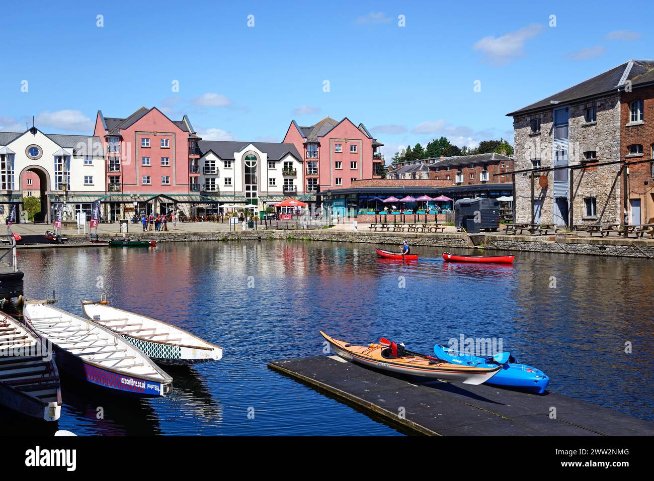 Kajakfahren auf dem Exeter Schiffskanal mit Cafés und Restaurants auf der Piazza Terracina auf der Rückseite und der Topsham Brauerei auf der rechten Seite, Exeter, Großbritannien. Stockfoto
