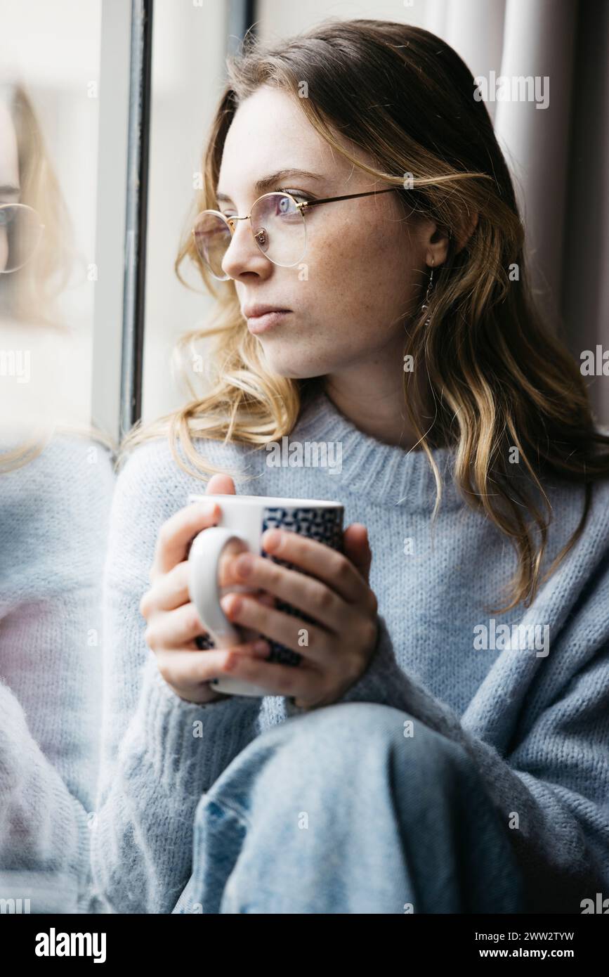 Nachdenkliche junge, schöne blonde Frau, die eine Tasse Tee an einem Fenster in ihrem Haus hält, was eine intime und gemütliche Atmosphäre schafft Stockfoto