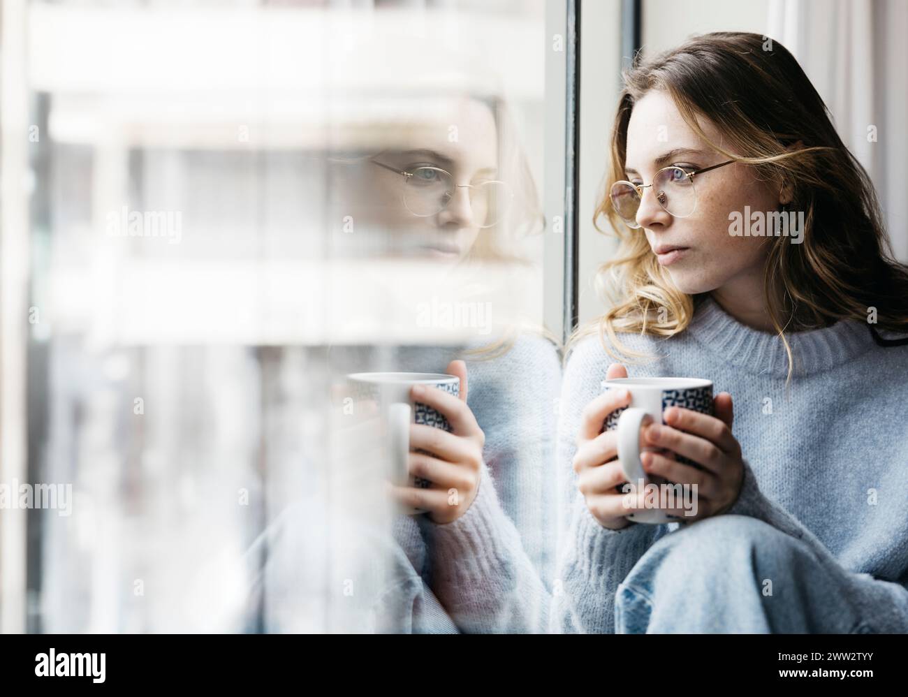 Nachdenkliche junge, schöne blonde Frau, die eine Tasse Tee an einem Fenster in ihrem Haus hält, was eine intime und gemütliche Atmosphäre schafft Stockfoto