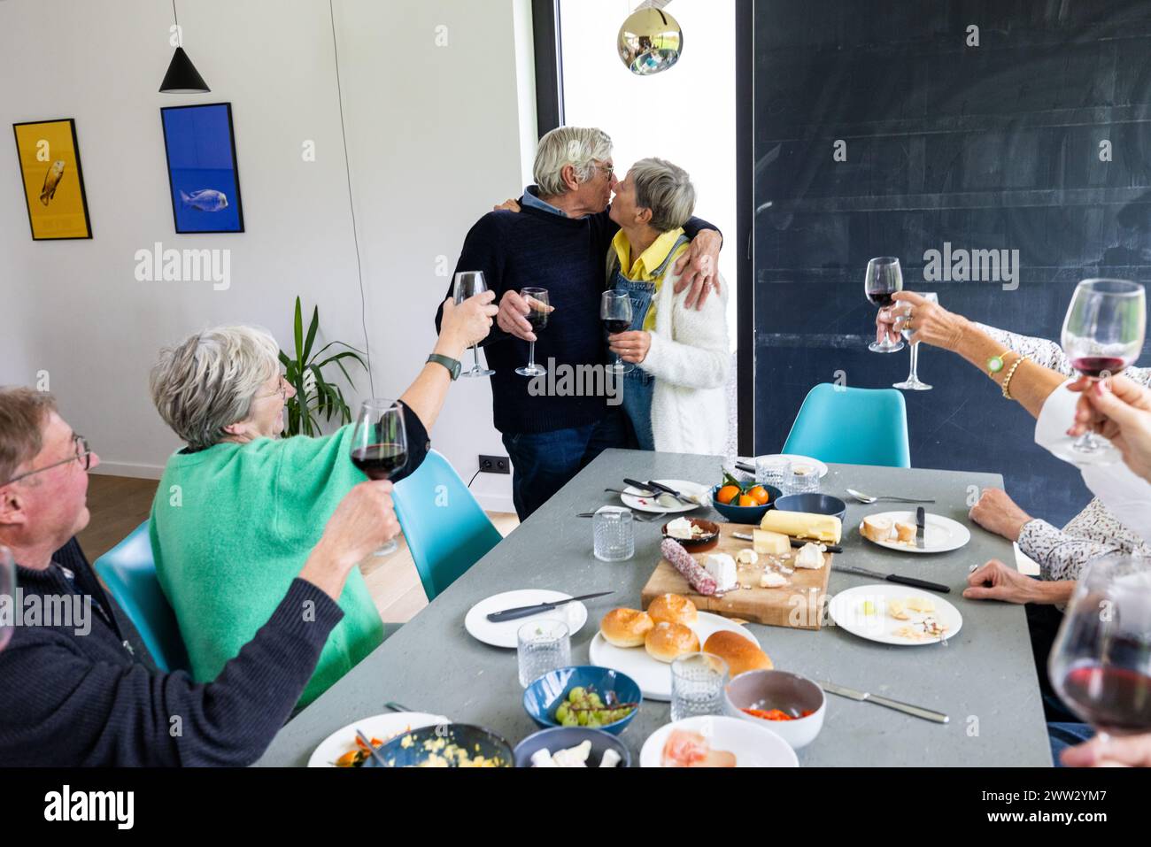 Ein intimes Familientreffen, bei dem ein älteres Paar einen liebevollen Kuss teilt, während andere ihre Gläser zum Toast heben. Die Inneneinrichtung verfügt über einen Esstisch, der mit einer Auswahl an Käse, Obst und Brot geschmückt ist, was eine gemütliche Atmosphäre der Feier schafft. Älteres Paar küsst sich bei Family Meeting Toast. Hochwertige Fotos Stockfoto